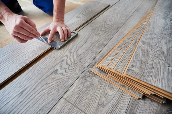 Close up of male worker using metal construction ruler and pen while drawing line on laminate flooring board. Man preparing laminate plank for floor installation in apartment under renovation.